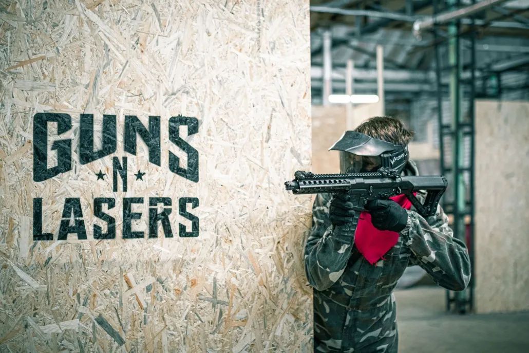 Uz OSB sienas GUNSnLASERS logo un airsoft spēlētājs ķiverē un ar ieroci tēmē iekštelpās.